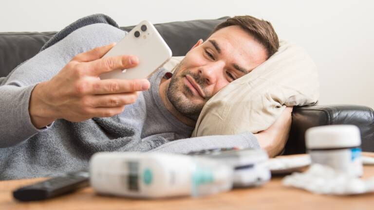 Ein Mann liegt auf einem Sofa und schaut aufs Smartphone, auf dem Tisch vor ihm liegen u. a. Medikamente und ein Fieberthermometer