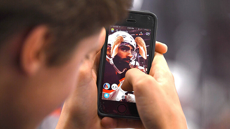 Ein Mann macht ein Foto von einem Eishockey-Spieler und nutzt einen Filter. Dieser zeigt am Helm des Spielers Hundeohren an.
