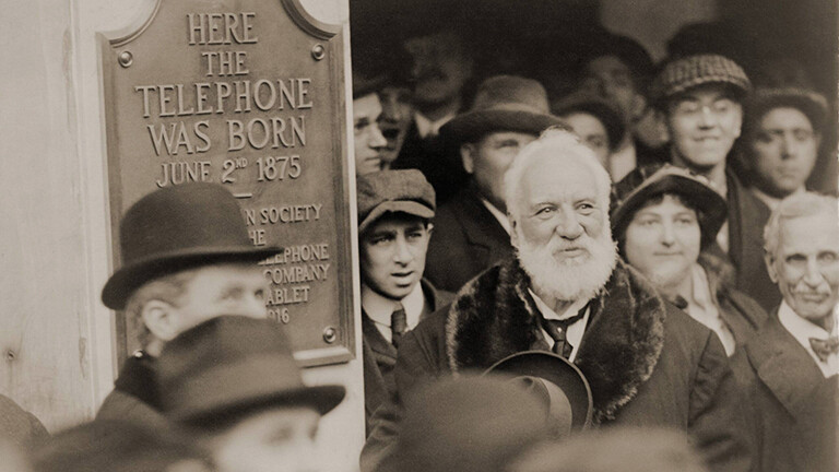 Schwarz-weiß Archifoto: Der Erfinder Graham Bell steht inmitten einer Menschengruppe. An der Wand neben ihm wurde ein Schild angebracht, das ihn als Erfinder des Telefons ehrt.