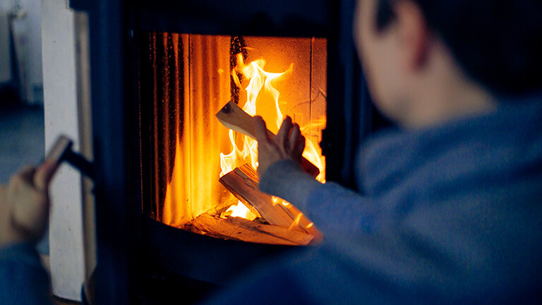 Eine Person sitzt vor einem Kaminofen, darin brennendes Holz. Der Mann hat die Kamintür geöffnet und legt Holz nach.