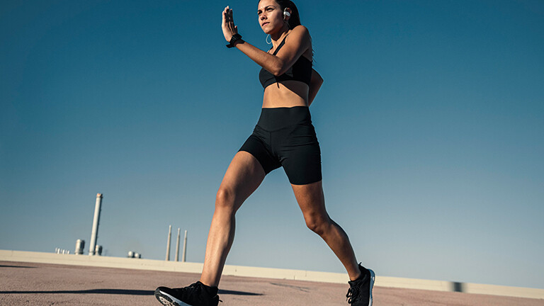 Eine junge Frau joggt draußen vor blauem Himmel. Sie wirkt sehr fit und dynamisch. Im Hintergrund sind Schornsteine zu sehen.