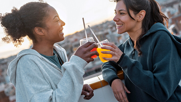 Zwei Frauen stehen auf einer Dachterrasse, es ist sommerlich. Sie lachen und stoßen mit zwei Getränken an.