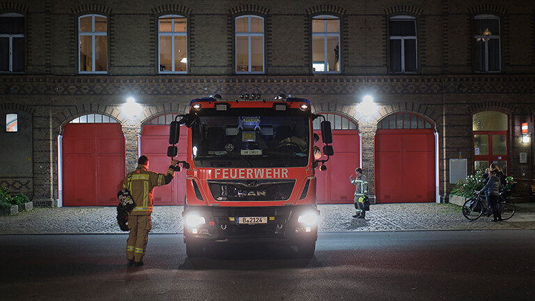 Vor einer Feuerwache steht ein Feuerwehrauto. Zwei Feuerwehrleute stehen am Auto. Es ist Nacht.