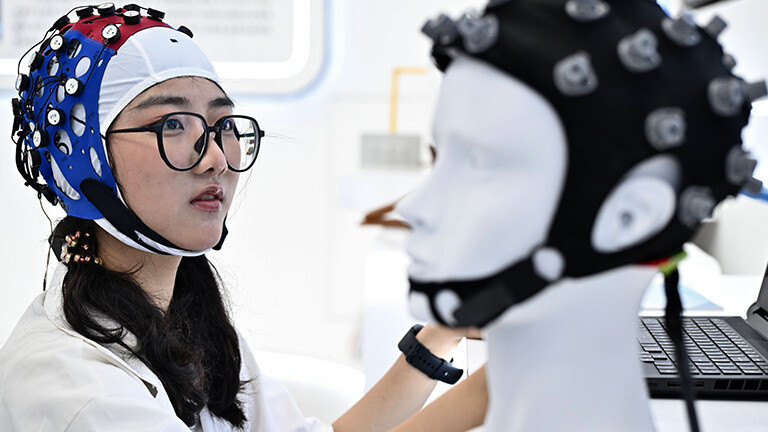 Bei einer Messe in China trägt eine Besucherin eine Art Mütze mit Elektroden, vor ihr steht ein Modellkopf, ebenfalls mit Mütze und Elektroden.
