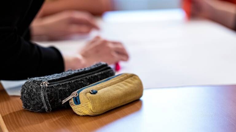 Zwei Federmäppchen liegen auf einem Tisch, im Hintergrund lehnt sich eine Person über den Tisch