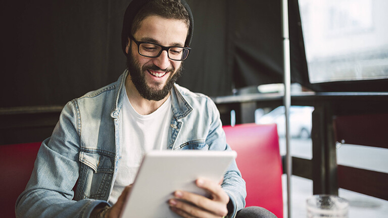 Ein Mann mit Brille und Mütze hält ein Tablet in der Hand und lächelt.
