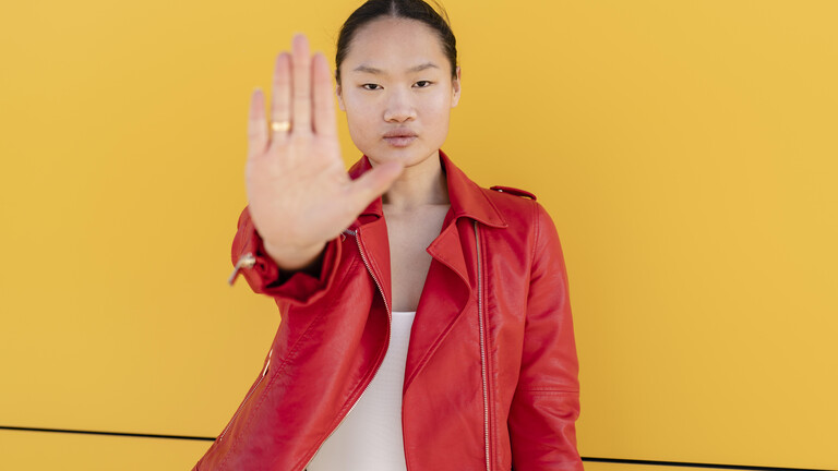Eine Frau steht vor einer gelben Wand. Sie trägt eine rote Lederjacke, schaut in die Kamera und hält ihre flache Hand hoch zu einer Stopp-Geste.
