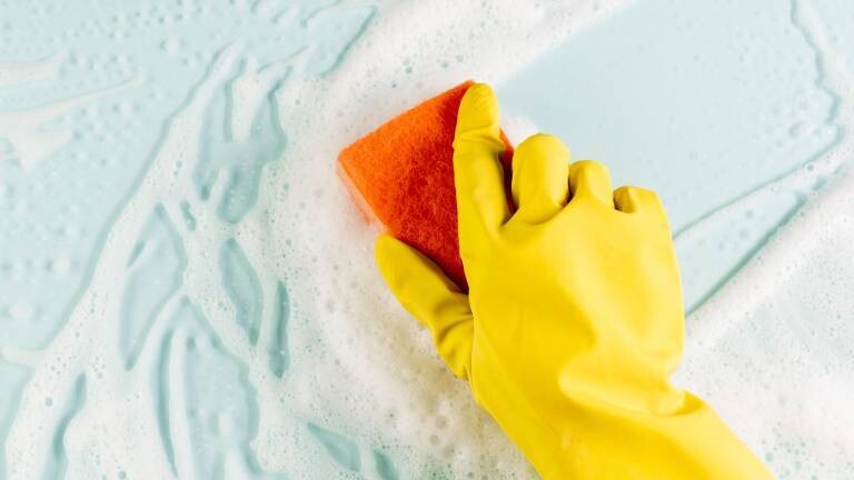 Hand mit gelben Putzhandschuh wischt mit Schwamm über weißen Schaum