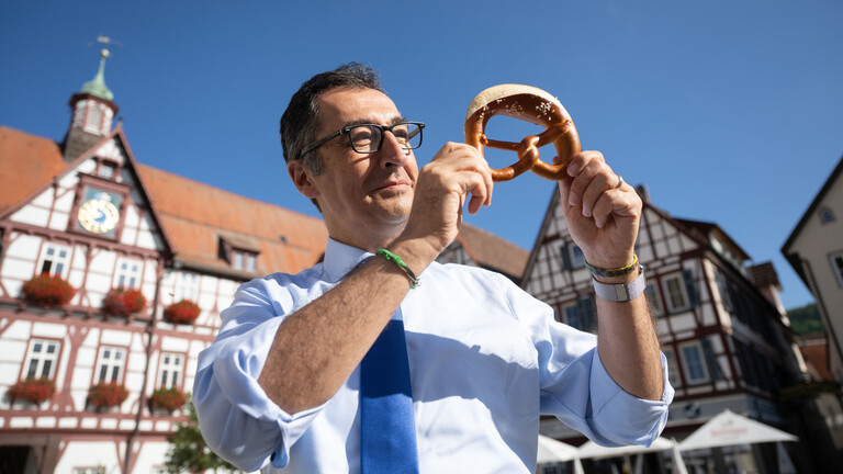 Bundesernährungsminister Cem Özdemir (Bündnis 90/Die Grünen) zeigt auf dem Marktplatz seines Geburtsorts nach dem Besuch einer Bäckerei eine Brezel.
