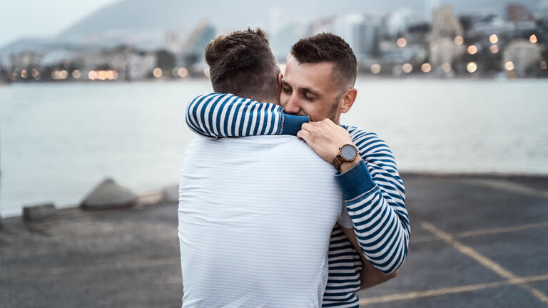 Ein Liebespaar umarmt sich. Im Hintergrund ein Hafenbecken und unscharf eine Stadt