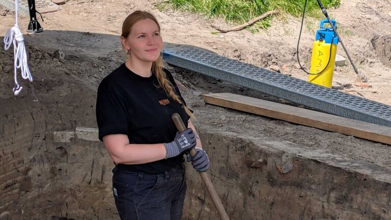 Archäologin Svea Weihmann steht in einem Ausgrabungsfeld, trägt Arbeitskleidung und schwarze Handschuhe. In den Händen hält sie ein Werkzeug.