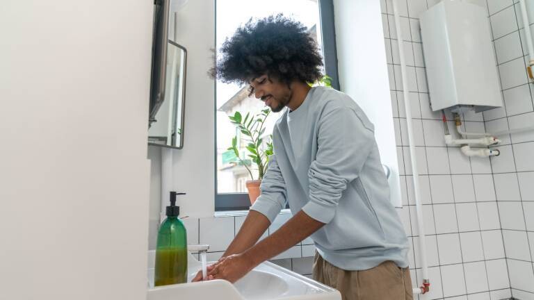 Ein Mann steht am Waschbecken im Bad und wäscht sich mit flüssiger Seife unter laufendem Wasser die Hände.