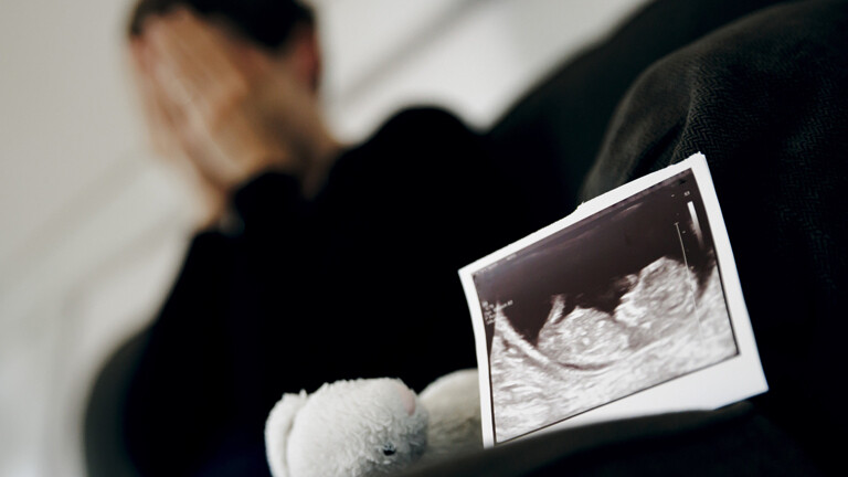 Gestellte Aufnahme zum Thema Fehlgeburt. Das Ultraschallbild eines ungeborenen Kindes steht neben einem Kuscheltier waehrend eine Frau im Hintergrund ihr Gesicht in ihren Haenden verbirgt