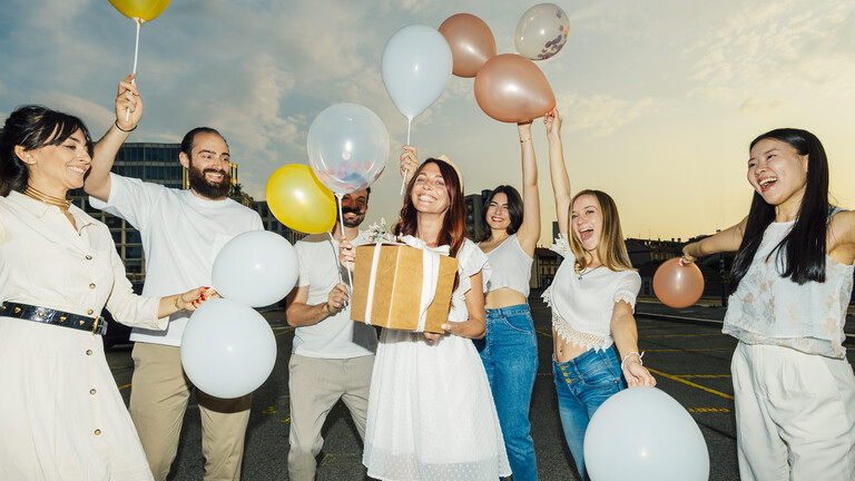 Leute lachen und halten Luftballons in den Händen, sie stehen um eine Frau herum, die ein Geschenk in den Händen hält