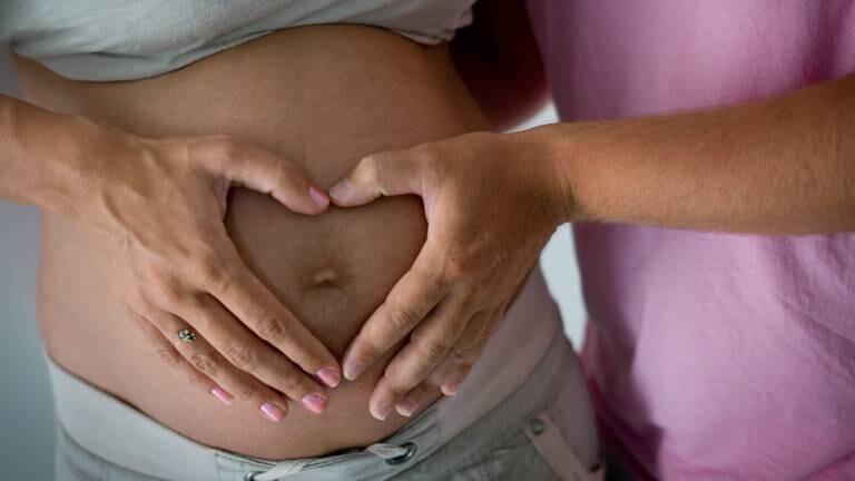 Die Hand einer Frau und die eines Mannes liegen in Herzform auf dem schwangeren Bauch der Frau