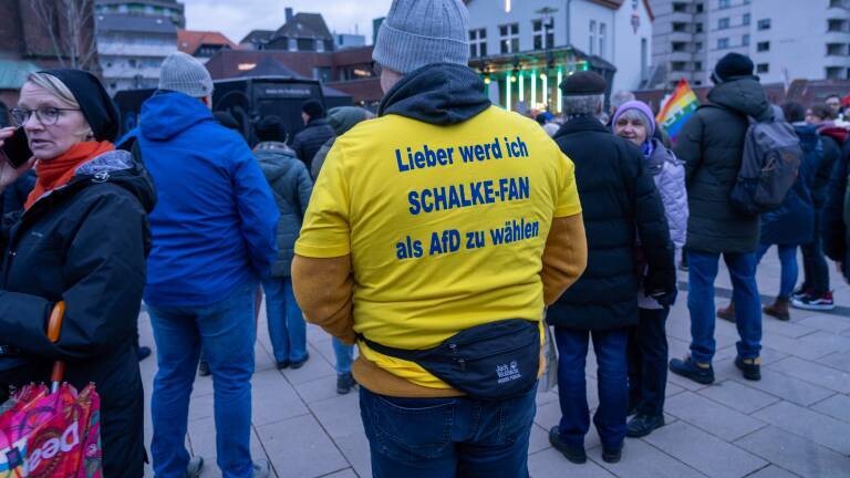 Ein Mann steht bei einer Demo gegen die AfD mit dem Rücken zur Kamera. Auf seinem gelben T-Shirt steht in Blau: „Lieber werde ich Schalke-Fan als AfD zu wählen“.
