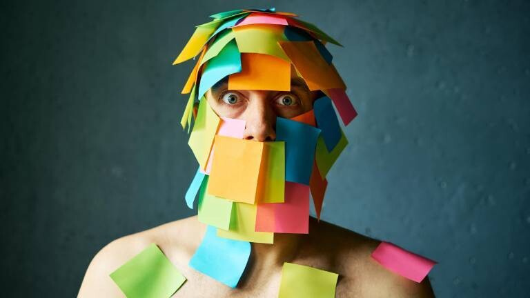 Ein Mann hat viele Post-its-Notes im Gesicht und auf den Schultern kleben und schaut gestresst