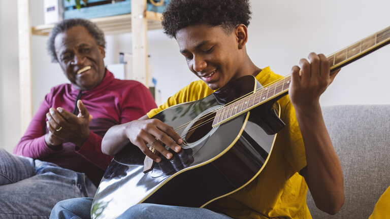 Lächelnder Junge spielt Gitarre bei seinem Großvater auf dem Sofa zu Hause
