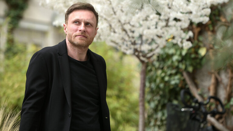 Der deutsche Autor Michael Nast steht draußen in einem Garten, er trägt ein schwarzes Jackett und Oberteil. Im Hintergrund blüht ein weißer Kirschbaum.