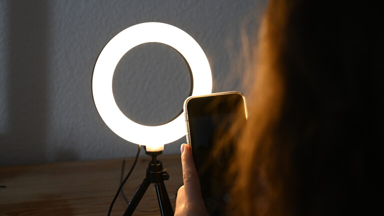 Eine Frau sitzt hinter einem Ringlicht und macht dabei Aufnahmen von sich.