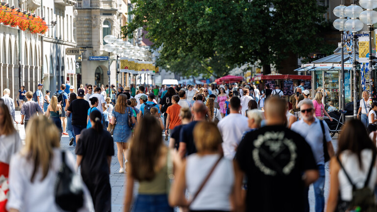 In München sind an einem Sommertag viele Menschen in der Innenstadt unterwegs. Sie gehen an Bekleidungsgeschäften vorbei.