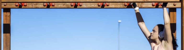 Porträt einer sportlichen Frau beim Klimmzug am Barren an einem sonnigen Tag mit Kopfhörern und Sportkleidung