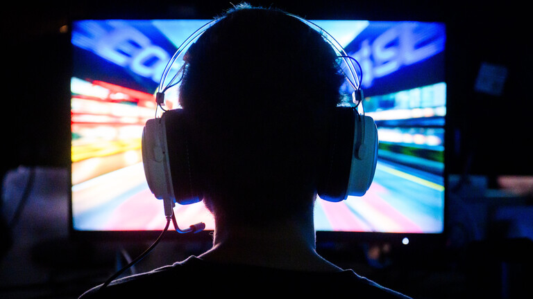 Ein Gamer spielt an ein Computerspiel. Wir sehen ihn von hinten. Er hat Kopfhörer mit Mikrofon auf.