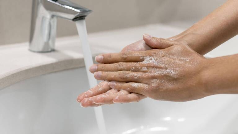 Eine Person wäscht ihre Hände mit Seife und Wasser an einem Waschbecken, an dessen Wasserhahn Wasser herausläuft.