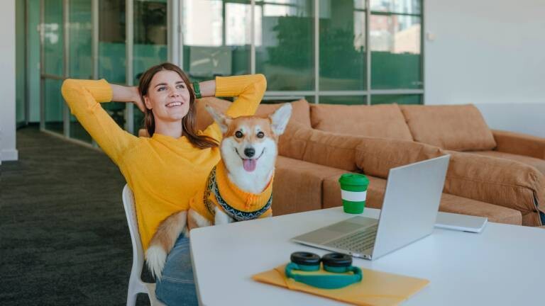 Eine Frau macht eine Pause während der Arbeit. Auf ihrem Schoß sitzt ihr Hund. Auf dem Schreibtisch stehen Laptop, Kaffee, Kopfhörer sowie PC-Zubehör.