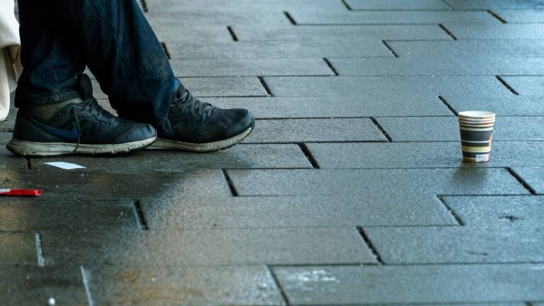 Schuhe eines Menschen, vor ihm steht ein Pappbecher