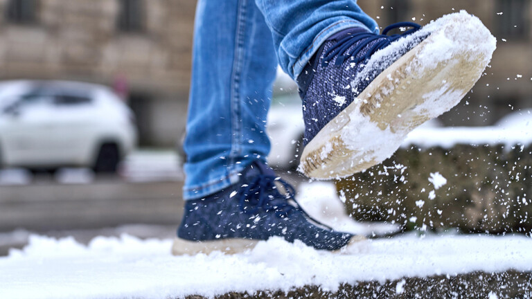 Ein Fuß einer Person tritt in den Schnee auf eine vereiste Treppe. Die rutschige Oberfläche und die Schneeschicht auf dem Gehweg symbolisieren die Gefahr von Glätte und Ausrutschen im Winter bei falschen Schuhwerk.