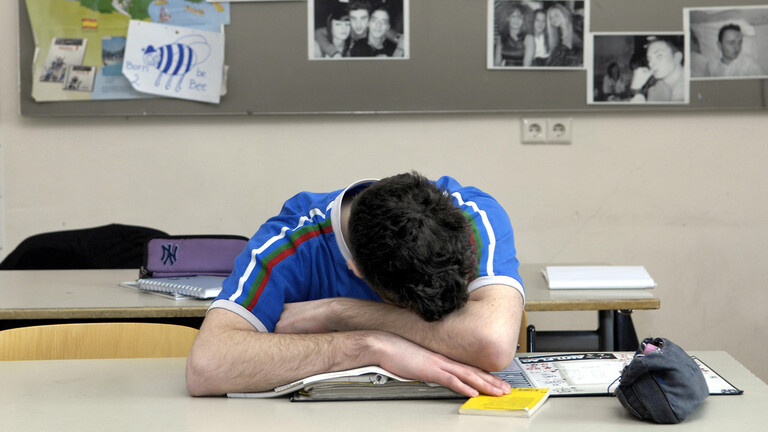 Ein junger Mann mit blauem Shirt und dunklen Haaren, der mit dem Kopf auf dem Tisch über einem Buch liegt.