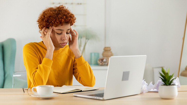 Übermüdete junge Frau beim Arbeiten am Laptop, sie hat Kopfschmerzen und trockene Augen und massiert sich die Schläfen, eine Kaffeetasse steht auf dem Tisch (Symbolbild)