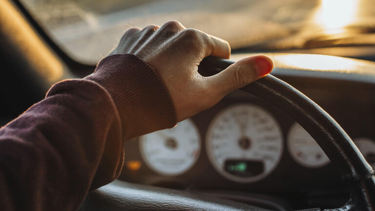 Eine Hand am Lenkrad in einem Auto mit Blick auf einen Sonnenuntergang draußen