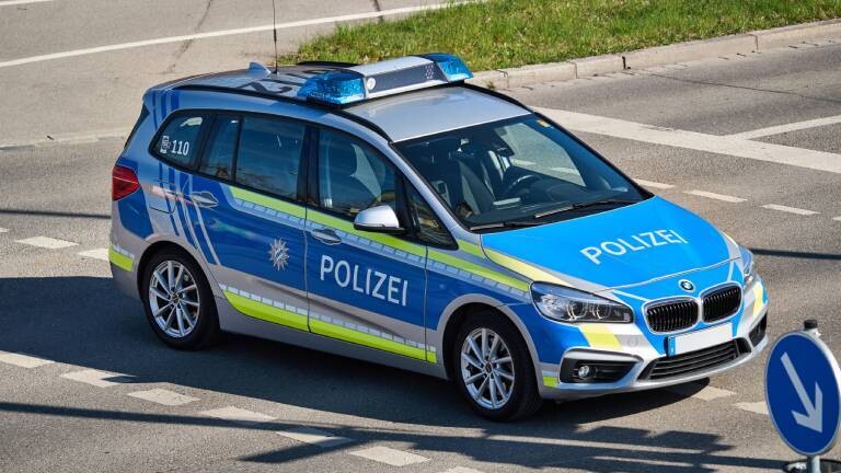 Ein silber-blaues Polizeiauto, ein Kombi mit neongelben Warnstreifen und dem großen Schriftzug "POLIZEI", steht schräg auf einer asphaltierten Straße. Das Blaulicht auf dem Dach ist ausgeschaltet. Im Vordergrund rechts ist ein blau-weißes Abbiege-Verkehrsschild zu sehen.