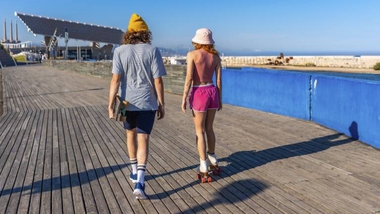 Ein junger Mann mit einem Skateboard spaziert neben einer Rollschuh fahrenden Frau auf einem Holzsteg