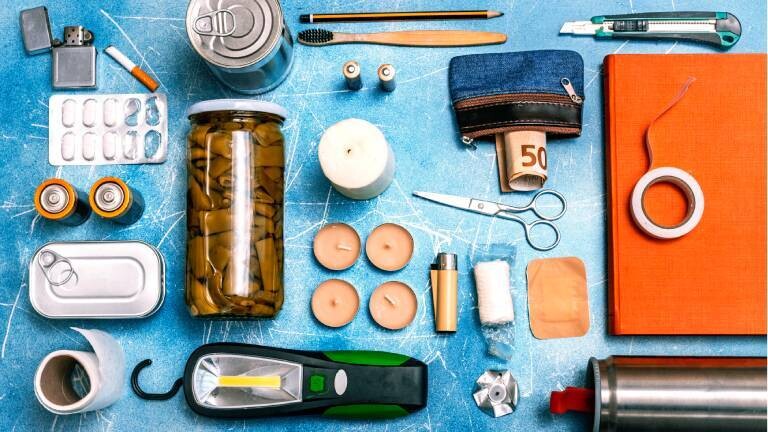 Survival-Essentials-Kit mit Kerzen, Feuerzeug, Batterien, Konserven und Solar-Taschenlampe vor blauem Hintergrund