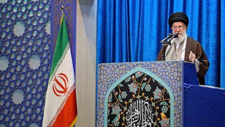Ayatalloh Ali Khamenei bei Rede vor blauem Samthintergrund neben iranischer Flagge