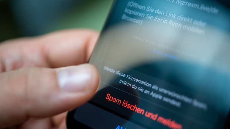 Ein Mann hält ein Smartphone in seiner Hand. Auf dem Bildschirm ist eine Spam-Nachricht geöffnet. Er klickt auf den Button "Spam löschen und melden".