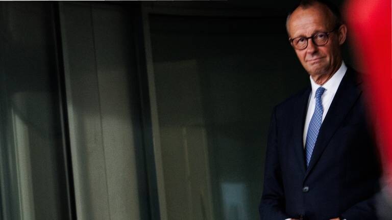 Bundeskanzler Friedrich Merz steht am Rand einer Bühne. Im Hintergrund ist ein dunkler Vorhangstoff zu sehen. Er trägt eine Brille, einen dunkelblauen Anzug, ein weißes Hemd und eine hellblau gemusterte Krawatte.