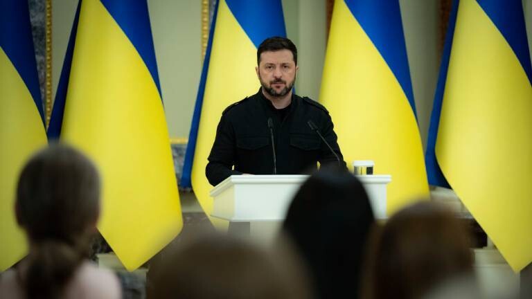Der ukrainische Präsident Volodymyr Selenskyj steht am Mikrofon, hinter stehen ukrainische Flaggen in einer langen Reihe