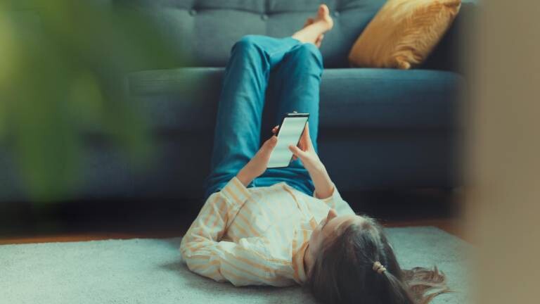 Frau liegt auf Boden, hat die Beine aufs Sofa gelegt und scrollt auf dem Handy