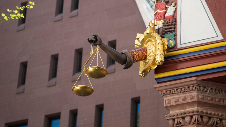 Ein ausgestreckter Arm aus Holz hält eine goldene Waage an einem rekonstruierten Fachwerkhaus