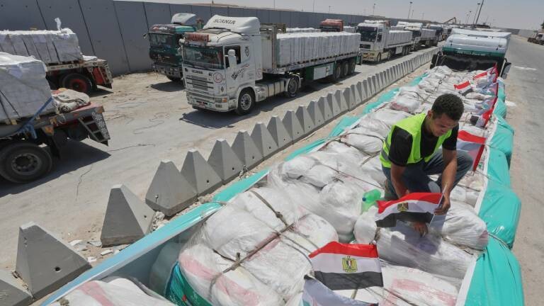 Mann mit gelber Warnweste befestigt ägyptische Flaggen auf Hilfslieferungen, die auf einem LKW liegen. Mehrere solcher LKWs stehen an der Grenze Gaza-Ägypten (06.08.2025)