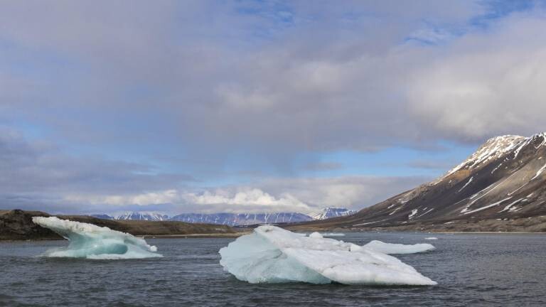 Eine Eisscholle treibt im Meer in der Bucht vor dem Gletscher Recherchebreen bei Spitzbergen, Svalbard. Der Himmel ist bewölkt bis heiter. Die Berge im Hintergrund sind nur wenig mit Schnee bedeckt.
