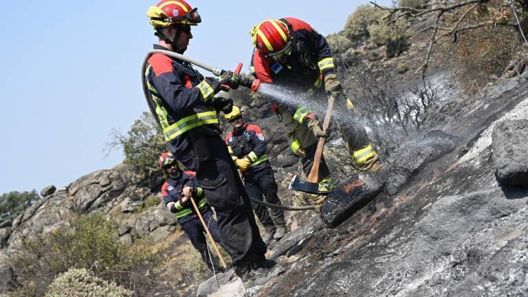 Deutsche Feuerwehrleute spritzen in der Region Extremadura Wasser auf einen bereits abgebrannten Bereich und suchen nach möglichen Glutnestern. Sie tragen Schutzkleidung und Helme. Die Pflanzen sind verkohlt und Felsen schwarz.