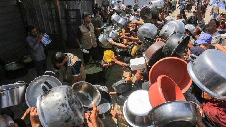 Palästinenser, darunter auch Kinder, die aufgrund der Blockade Israels und der anhaltenden Angriffe auf den Gazastreifen Schwierigkeiten haben, sich mit Lebensmitteln zu versorgen, stehen am 21. August 2025 in Khan Yunis im Gazastreifen in der Region Al-Mawasi Schlange, um warme Mahlzeiten zu erhalten, die von einer Wohltätigkeitsorganisation verteilt werden. Sie halten Töpfe und Schüsseln hoch über ihre Köpfe.