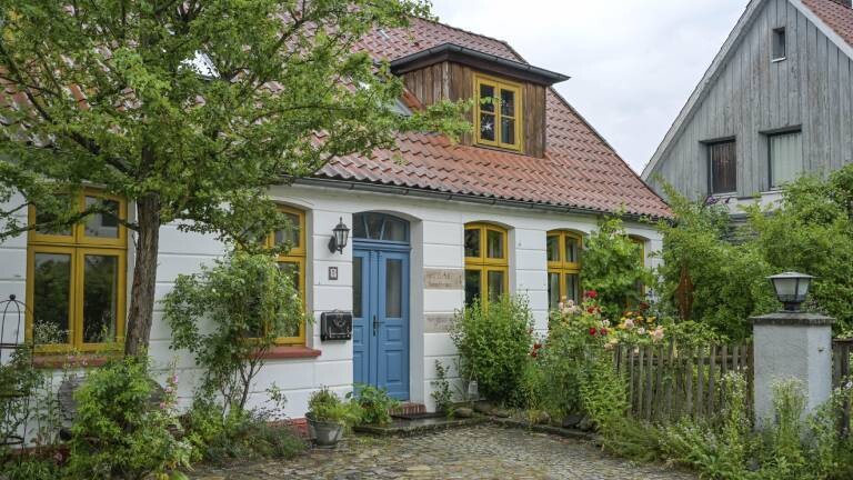 Ein Wohnhaus in einem ländlichen Stil von vor 100 Jahren. Vor dem Haus ist ein kleiner Garten mit Blumen zu sehen.