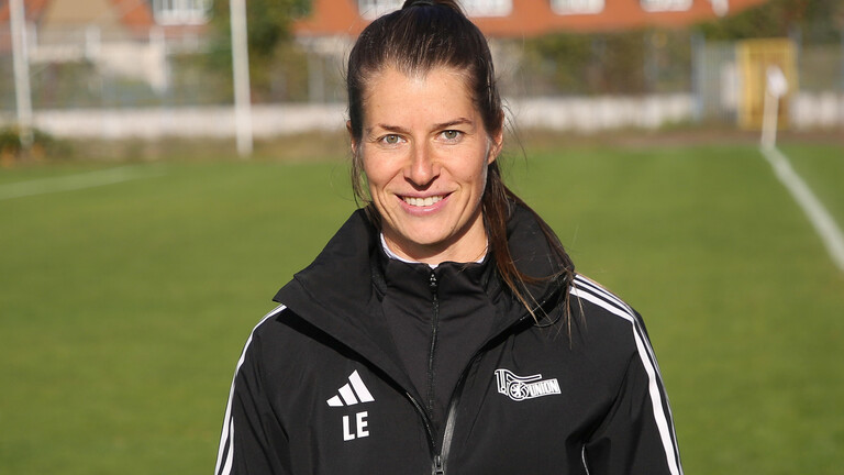 Marie-Louise Eta steht in einer Trainingsjacke auf einem Fußballplatz und schaut direkt in die Kamera.