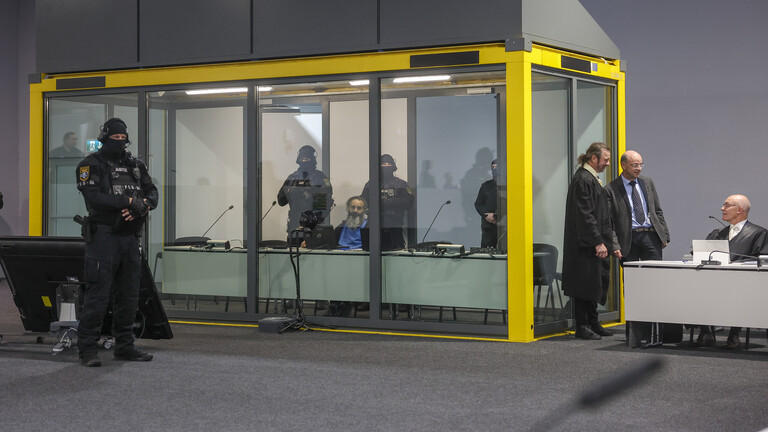 Hinter einem Glaskasten sitzt der Angeklagte, um ihn Polizisten